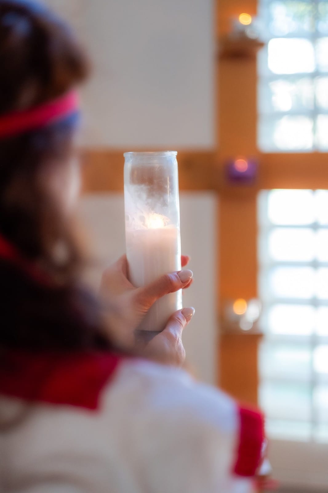 Marilyn holding a lit white candle in a glass vessel before a warm wooden cross