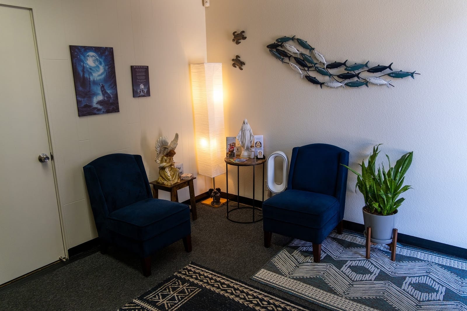 The waiting room with two navy blue velvet chairs, floor lamp, angel and Mary statues, and fish wall art